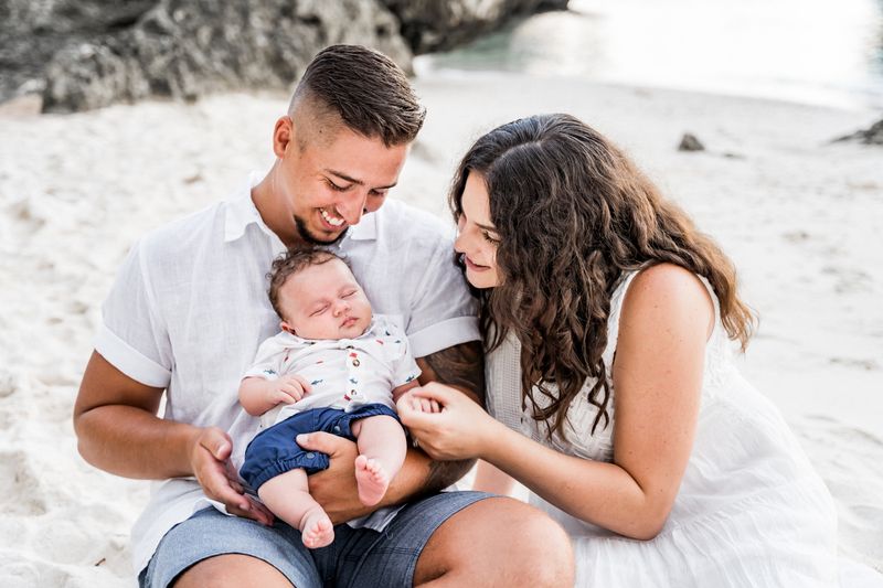 family of three with a newborn, sitting on a beach and looking at the sleeping baby