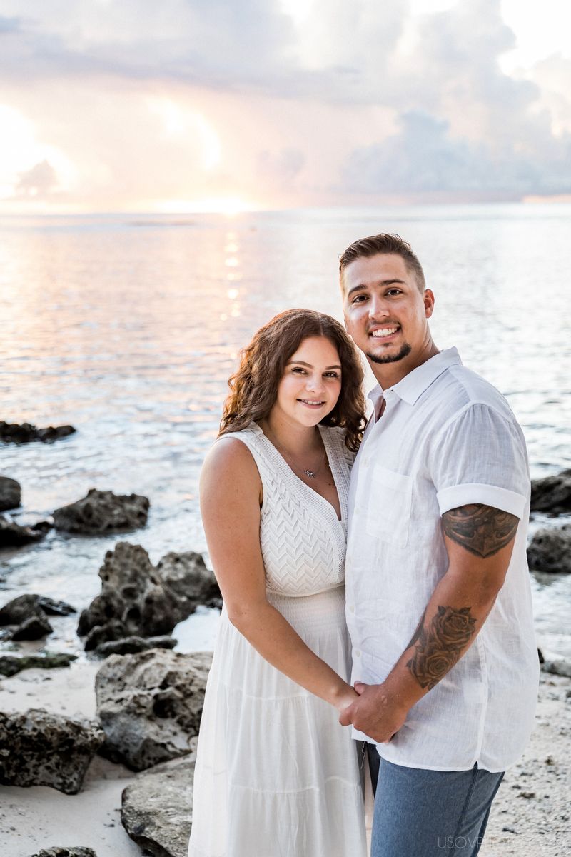 couple in white outfits on a beach, water reflecting pink sunset on the backgrounf