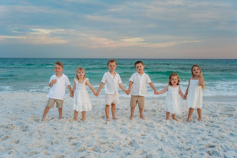 Six children holding hands in matching white and beige outfits on the sandy beach in Destin, Florida, at sunset