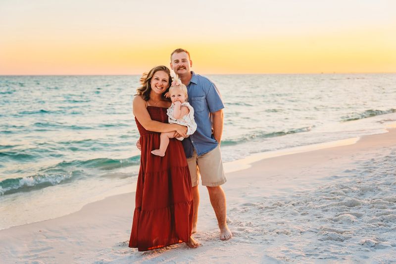 Family of three wearing a red maxi dress, blue shirt, and white baby outfit for sunset beach portraits in Destin, Florida
